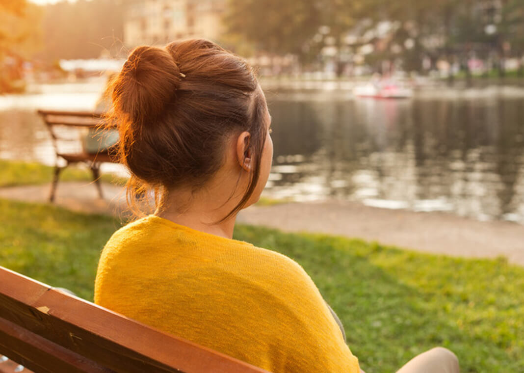 young woman with hair in bun sitting on a bench and looking at a body of water - SuzanaMarinkovic/iStock/Getty Images